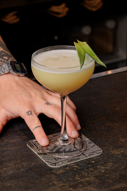 A hand placing a citrus-based Salford Spiced Rum cocktail in a coupe glass onto a coaster.